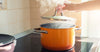 Hand lifting lid off steaming orange pot on stove